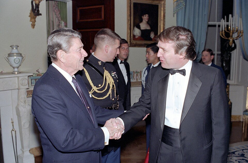 Ronald Reagan shaking hands with Donald Trump, who wears a dark formal jacket and bow tie at a 1987 White House reception